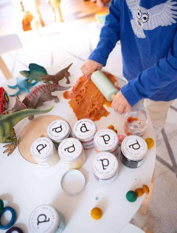 I child using a rolling pin to smooth out some orange play dough on a table surrounded by plastic dinosaurs