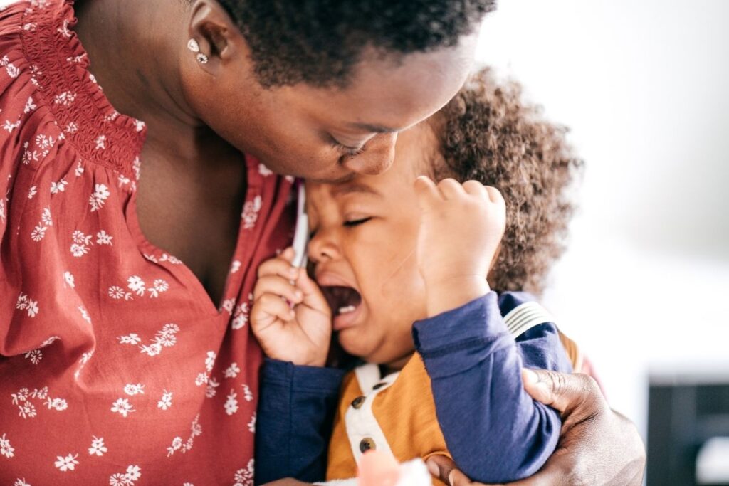 Black mother and child. Mother is kissing the forehead of the toddler. Toddler is exhibiting tricky toddler behavior.